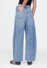 Pantalones vaqueros azules de pierna ancha con acabado desteñido, con dos bolsillos traseros y un dobladillo sin rematar. Combinados con zapatillas negras.