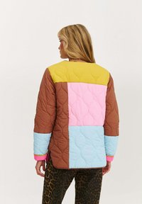 Žena s dlhými blond vlasmi, oblečená v prešívanom farebne rozdelenom veste v žltej, hnedej, ružovej a modrej farbe, v kombinácii s nohavicami so zvieracím vzorom.