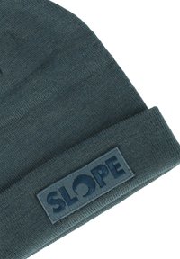 Mörkgrön stickad mössa med en vikta cuffs som har en rektangulär patch med texten "SLOPE" i upphöjda blå bokstäver.