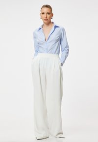 Camisa de botones a rayas azul claro con cuello, combinada con pantalones anchos blancos con plisados. El modelo está de pie con las manos en los bolsillos.