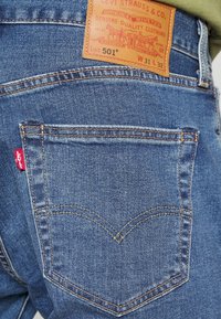 Jean en denim bleu Levi's 501 montrant la poche arrière avec la couture signature, l'étiquette rouge Levi's et un patch de marque en cuir marron avec les détails de la taille.