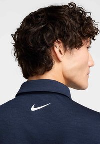 Tamsiai mėlynas polo marškinėliai su tekstūruota medžiaga, ant nugaros esantis baltas Nike logotipas. Apykaklė tradicinė ir standi.