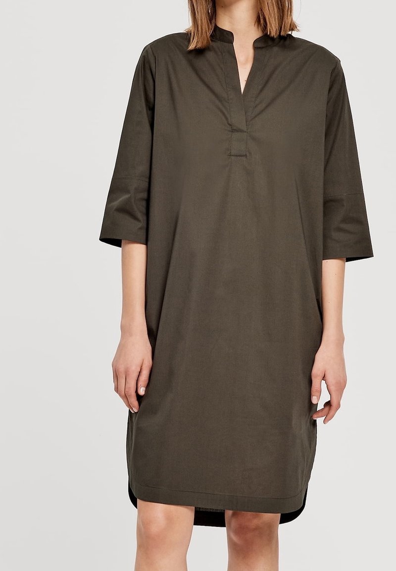 Robe ample vert olive avec manches trois quarts, col montant et fente en V au niveau du décolleté, portée par une personne cadrée au niveau du cou et des genoux.