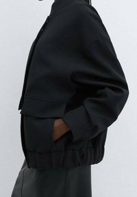 Veste noire cropped avec un col haut, dotée d'une fermeture éclair, de manches larges et d'un ourlet froncé pour une silhouette structurée.