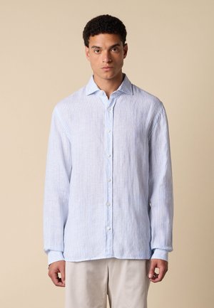 Giovane uomo rivolto in avanti che indossa una camicia a righe azzurro chiaro a maniche lunghe con bottoni e pantaloni beige su uno sfondo beige uniforme.