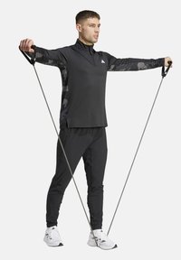 Top atlético negro de manga larga con un patrón de camuflaje; combinado con pantalones negros a juego y zapatillas deportivas blancas, sosteniendo bandas de resistencia.