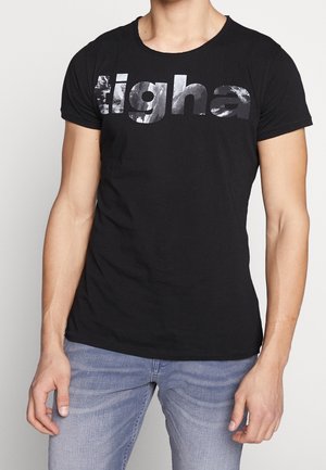 Mann trägt ein schwarzes, eng anliegendes T-Shirt mit großem metallisch-silbernem "tigha" Schriftzug auf der Brust und hellblauen Jeans.