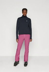 Tengerészkék hosszú ujjú felső magas gallérral és kis logóval, rózsaszín sínadrággal és fekete síbakancsokkal, semleges háttér előtt.