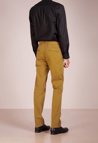 Mężczyzna w musztardowych prostych spodniach, czarnej koszuli eleganckiej i czarnych skórzanych butach, stoi tyłem na neutralnym tle.