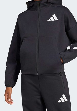 Persoon draagt een zwart hoodie met rits en een broek met wit Adidas-logo op de borst en bovenbeen, hand in broekzak.