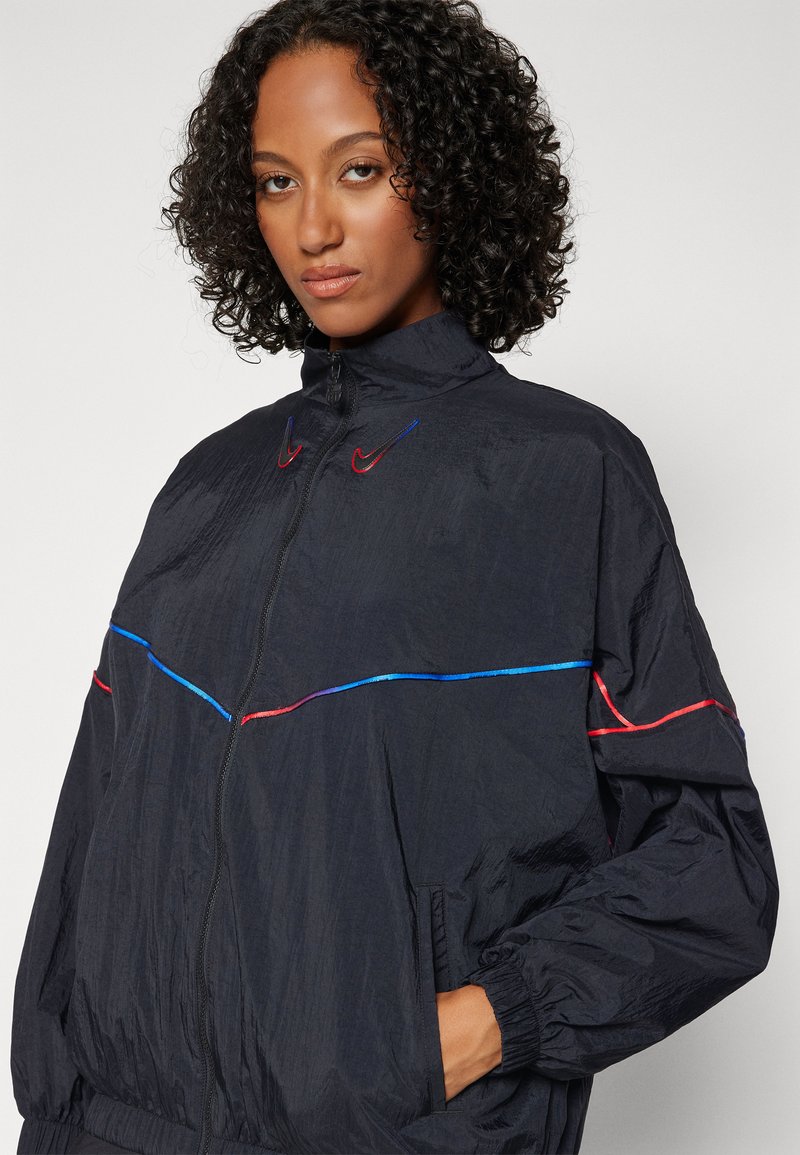 Schwarze Windbreakerjacke mit hohem Kragen, Reißverschluss vorne und mehrfarbigen Streifen. Das Material hat ein leichtes, strukturiertes Finish.