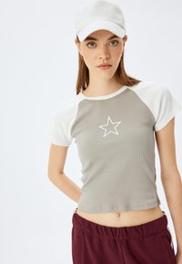 Grå ribbet crop top med hvide raglanærmer og et hvidt stjernemotiv på brystet. Modellen har maroon sweatpants og en lys grå cap på.