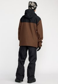 Veste bicolore marron et noire résistente aux intempéries avec capuche, associée à un pantalon noir. Un tissu texturé et des gants complètent le look.