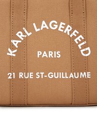 Tan fabric bag featuring white printed text: "KARL LAGERFELD PARIS 21 RUE ST-GUILLAUME." The design includes vertical seam lines.