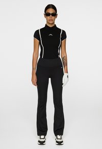 Donna che indossa un polo atletico nero, pantaloni neri, guanto da golf bianco sulla mano sinistra, occhiali da sole neri e sneakers bianco e nere, in piedi su uno sfondo bianco.