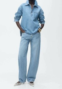 Jasnoniebieska koszula dżinsowa z kołnierzykiem i przednią kieszenią, zestawiona z szerokimi jasnoniebieskimi dżinsami. Białe buty na szpilce z wzorem w laserowym cięciu.