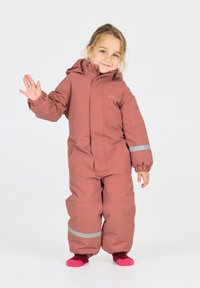 Roze waterdichte overall met capuchon, elastische manchetten en reflecterende strips, gedragen over roze sokken. Kind staat te zwaaien.