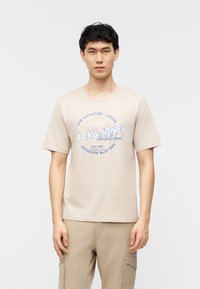 Νεαρός άνδρας με σκούρα μαλλιά, που φοράει μπεζ T-shirt με σχέδιο και ταιριαστό παντελόνι cargo, στέκεται μπροστά σε λευκό φόντο.