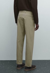 Man som bär beige byxor, mörkbrun långärmad skjorta och mörkbruna skor, står mot en ljus vägg med ett fläckigt golv.