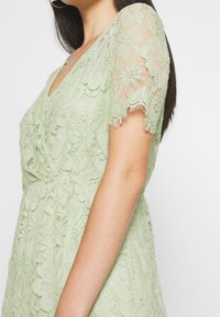 Robe en dentelle de couleur vert clair, avec des motifs floraux, des manches courtes et un col en V. Le tissu présente un design texturé et complexe.