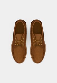 Zapatos de ante marrón con puntera redonda, dos ojales dorados para los cordones y costura tradicional a lo largo de los laterales. Suela plana de goma beige.