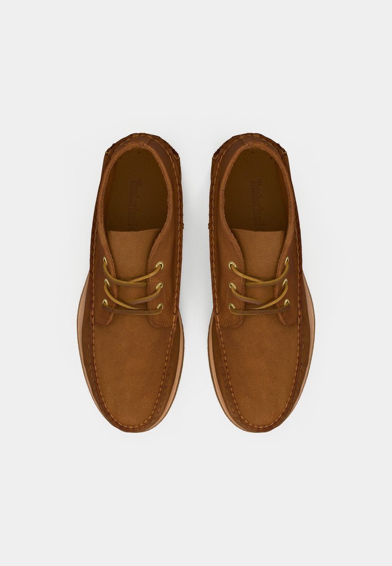 Scarpe in suede marrone con punta tonda, due occhielli dorati per i lacci e cuciture tradizionali lungo i lati. Suola piatta in gomma beige.