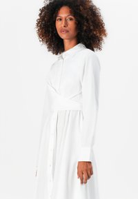 Abito bianco button-up con colletto, maniche lunghe e dettaglio avvolgente in vita. Presenta una gonna arricciata e una texture di tessuto liscia.