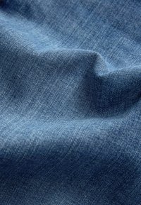 Tissu en denim bleu avec une texture de tissage visible et de petits plis créant des ombres et des reflets sur la surface.