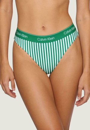 Parte inferiore del bikini a righe verticali verdi e bianche con fascia con logo Calvin Klein, indossata da una persona con pelle abbronzata e braccia rilassate.