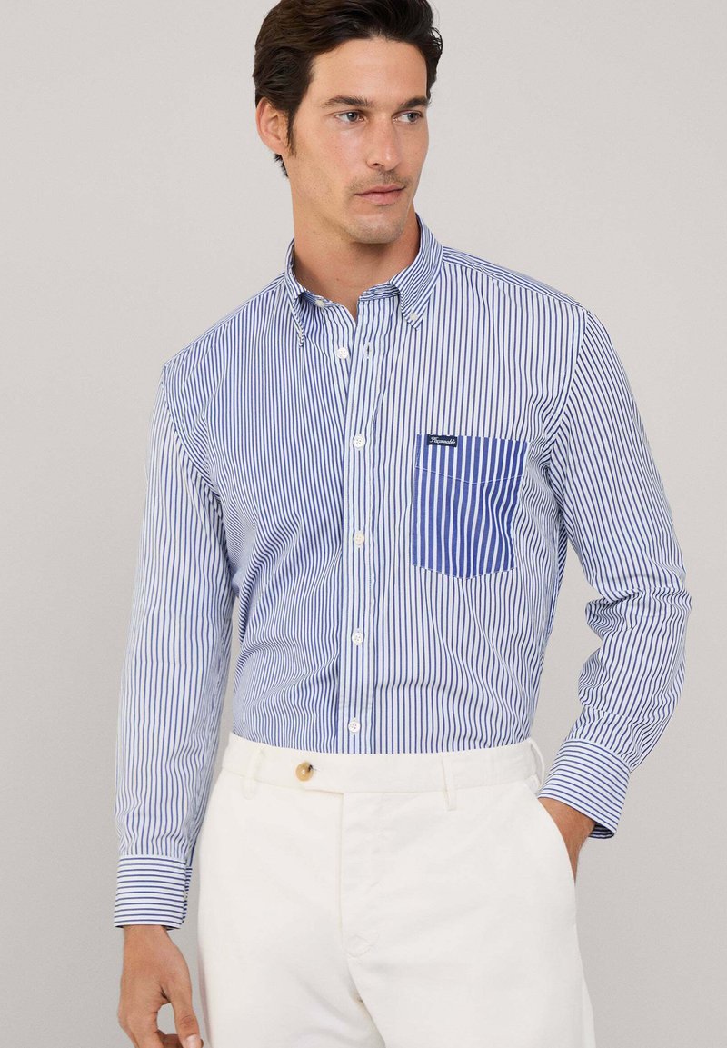 Camicia a righe blu e bianca con taschino sul petto con dettagli blu. Maniche lunghe con polsini abbottonati. Indossata con pantaloni bianchi.
