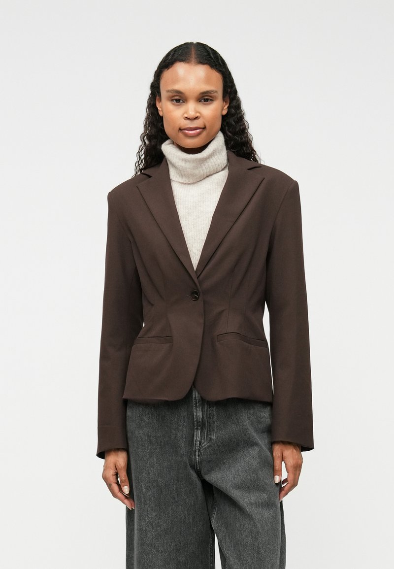 Brūns pielāgots žakets ar iegrieztiem uzšuvēm, ar vienu pogu aizdari, div�ām priekšējām kabatām un ciešu piegulšanu, valkāts virs gaiši pelēka augsta apkakle.