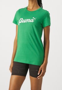 Žena v zeleném tričku Puma a černých sportovních šortkách s potiskem, stojící na bílém pozadí.
