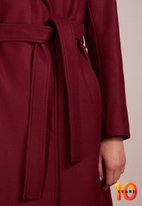 Abrigo burdeos envuelto de tejido texturizado con un cinturón anudado en la cintura y un suave cuello drapeado.
