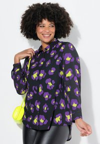 Sort skjorte med knapper og blomsterprint i lilla og neon grønne nuancer; har lange ærmer og en afslappet pasform, parret med en lille gul taske.