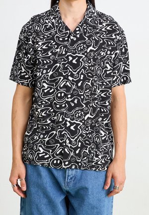 Man wearing black short-sleeve button-up shirt with white abstract and smiley face pattern, paired with blue jeans and silver rings on fingers.