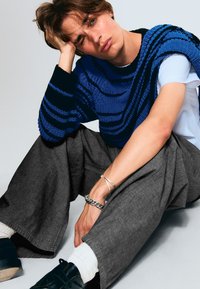 Pull en tricot à rayures bleu et noir, superposé à un t-shirt blanc. Pantalon gris ample et chaussures noires, avec des bijoux en argent au poignet.