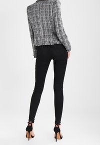 Žena v černobílé strukturované bundě, černých skinny džínách a černých sandálech na vysokém podpatku, stojí s otočením zády k divákovi.