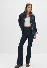 Kestenjak od trapera tamnoplave boje s velikim džepovima i zatvaranjem na dugmad, uparen s bijelim crop topom i tamnim bootcut trapericama s kontrastnim šavovima.