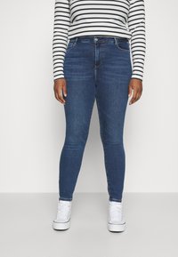 Tmavě modré skinny jeans vyrobené z denimu s hladkou texturou, se zapínáním na předním knoflíku a zipu. Nosené s proužkovanou košilí s dlouhým rukávem.