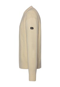 Beżowy sweter dzianinowy z teksturowanym wzorem, ściągacze na mankietach i u dołu, z czarną łatą z logo na lewym rękawie. Prosty okrągły dekolt.