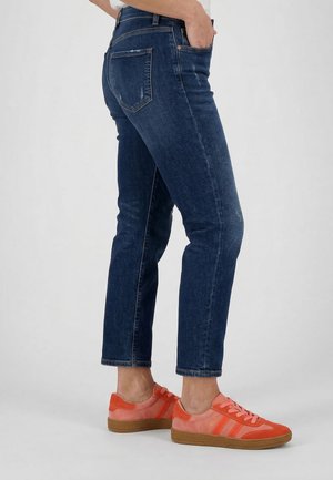 Person iført mørkeblå slim fit jeans og koral-pink sneakers med gummisål, stående sideværts mod en ensfarvet baggrund.