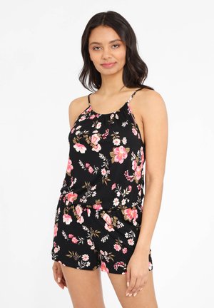 Junge Frau trägt schwarzen floralen ärmellosen Top und passende Shorts, steht vor weißem Hintergrund mit leichtem Lächeln.