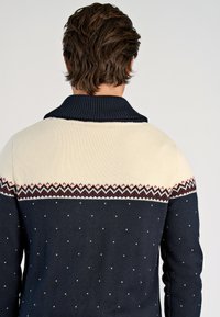 Marineblauwe en crèmekleurige gebreide trui met een geribbelde hals, voorzien van een geometrisch patroon op de borst en kleine witte accenten verspreid over de trui.