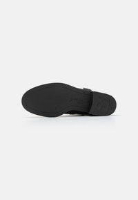 Suela de zapato negra con un acabado suave y mate. El diseño presenta un ligero arco, un logotipo en relieve y un tacón bajo y redondeado.