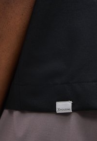 Κοντινό πλάνο της ραφής μαύρου ενδύματος με λευκή ετικέτα της μάρκας Houdini ραμμένη στο κάτω άκρο.