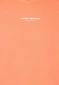 Kortärmad orange T-shirt med rund halsringning. Har vit text: "© WORST BEHAVIOR" och "PREMIUM 120Z 340G/M²" i mitten.