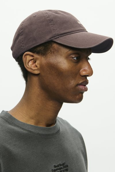 Mörkgrå keps i bomull med böjd skärm. Bärs tillsammans med en grå sweatshirt som har en rund halsringning och subtil broderad text.