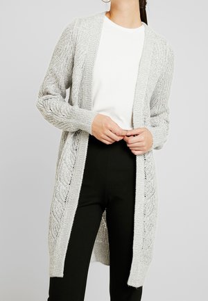Femme portant un cardigan en maille gris clair ouvert sur une chemise blanche et un pantalon noir, debout, les mains jointes.