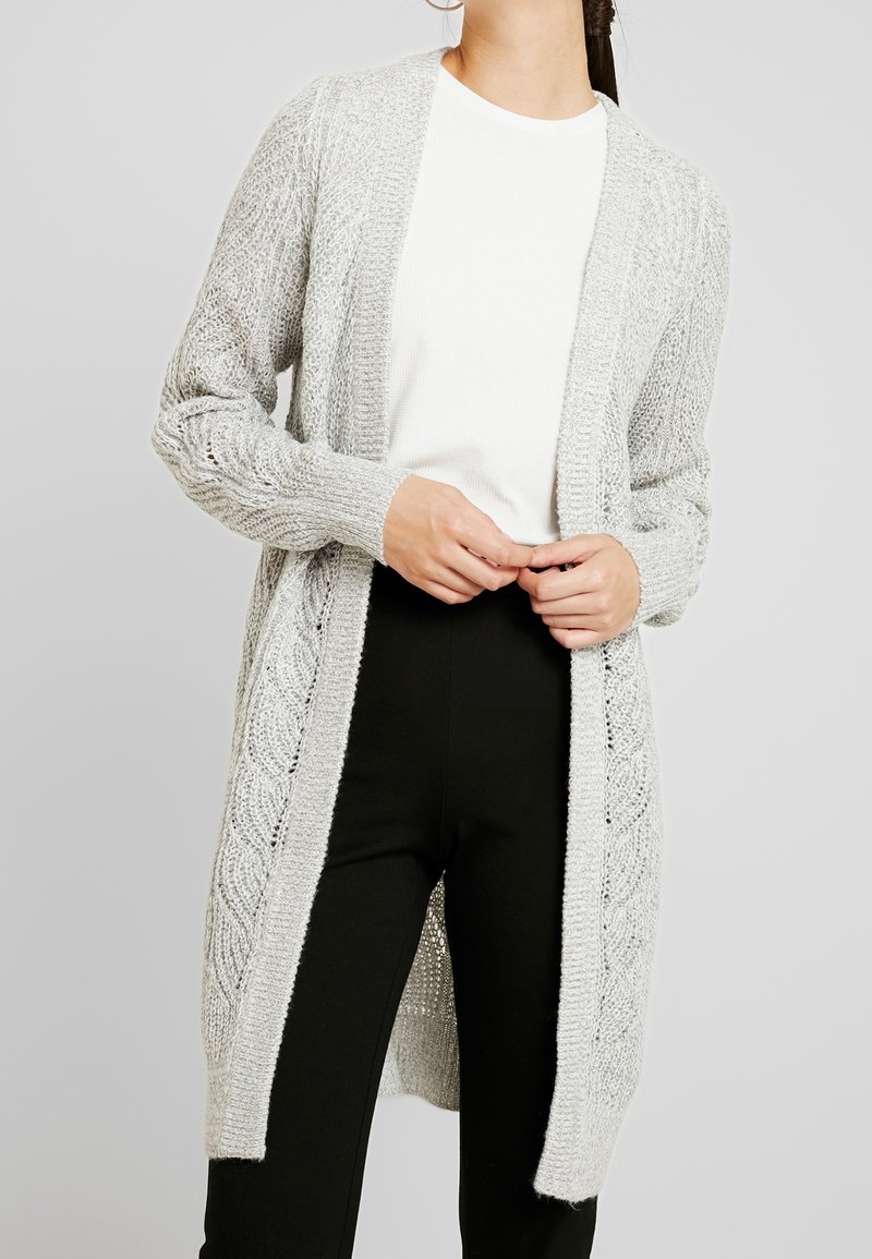 Femme portant un cardigan en maille gris clair ouvert sur une chemise blanche et un pantalon noir, debout, les mains jointes.