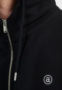 Schwarzer Kapuzenpullover mit Reißverschluss, silbernem Reißverschluss, strukturierten Kordeln und einem runden Logo-Patch mit einem Kleinbuchstaben ‚a‘ auf der Brust.